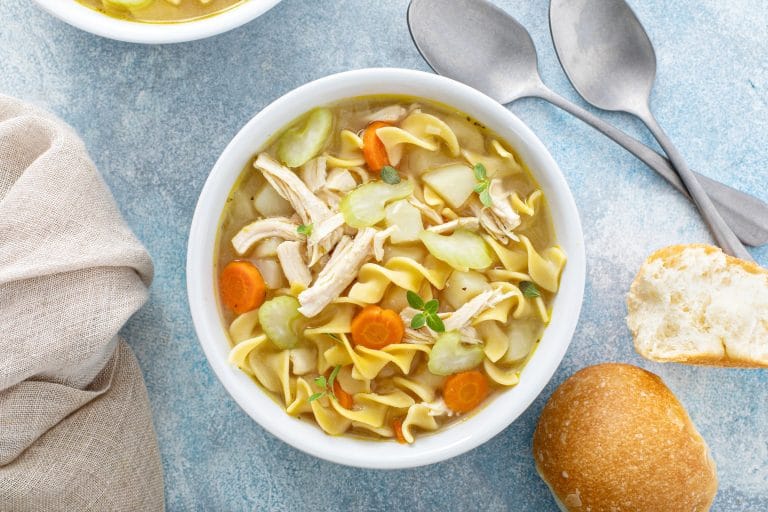 Soupe au poulet au aux nouilles avec carottes, céleri en morceaux servie dans un bol blanc avec du pain.