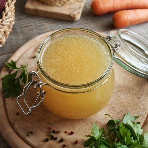 Bouillon de poulet maison fait à l'autocuiseur Instant Pot versé dand un pot mason sur une planche de bois avec des légumes autour.