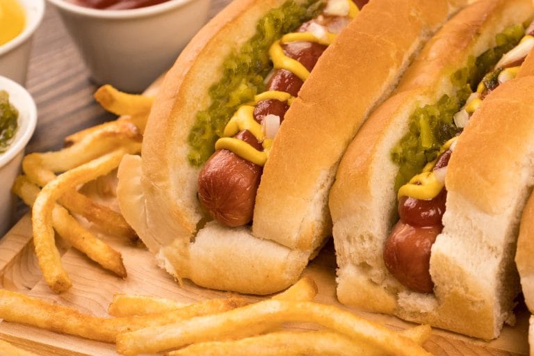 2 hot-dogs sur une planche de bois servis avec des frites, ketchup, moutarde, relish et oignons hachés
