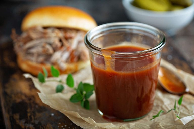 Sauce barbecue style Kansas City dans un pot avec un sandwich au porc effiloché et un bol de cornichons en arrière plan